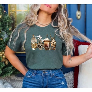 K&ouml;nnte beinhalten: Ein gr&uuml;nes T-Shirt mit einem Aufdruck von vier Kaffeegetr&auml;nken mit Totenk&ouml;pfen und Knochen. Die Getr&auml;nke sind ein Latte, ein Cappuccino, ein Mocha und ein Frappuccino.
