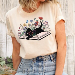 May include: A cream colored t-shirt with a black cat reading a book surrounded by colorful flowers. The cat is looking at the book and the flowers are blooming around it.