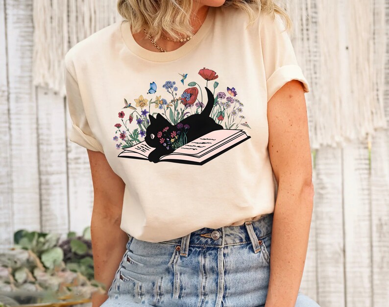 May include: A cream colored t-shirt with a black cat reading a book surrounded by colorful flowers. The cat is looking at the book and the flowers are blooming around it.