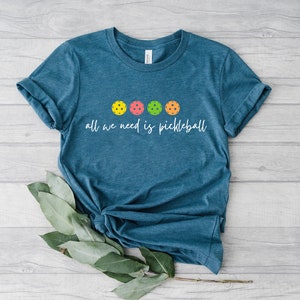 May include: A teal blue t-shirt with the text "all we need is pickleball" and five colorful pickleball balls.