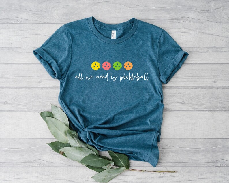 May include: A teal blue t-shirt with the text "all we need is pickleball" and five colorful pickleball balls.