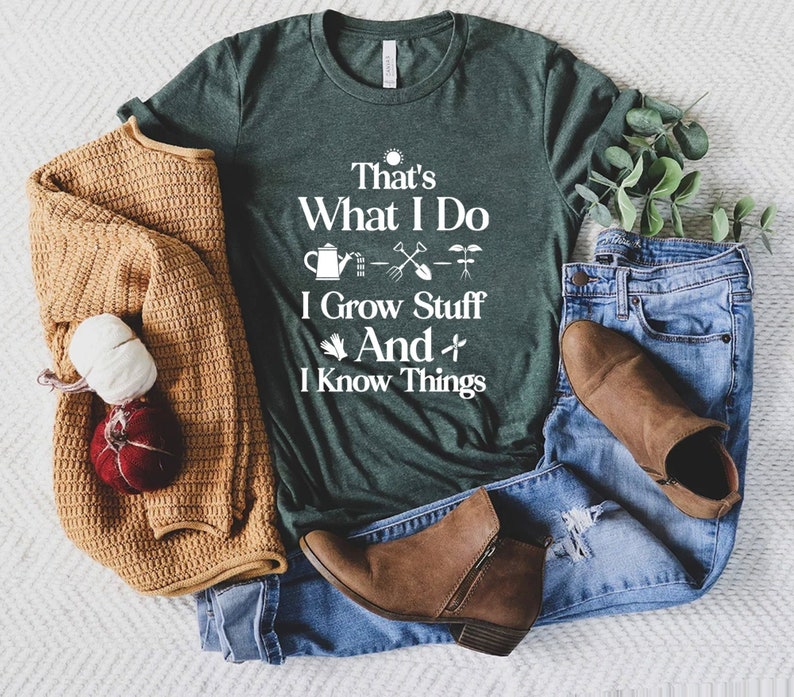 May include: A dark green t-shirt with white text that reads "That's What I Do I Grow Stuff And I Know Things". The text is accompanied by a sun, watering can, shovel, and plant icons. The t-shirt is laid out on a white surface with a brown sweater, blue jeans, and brown boots.