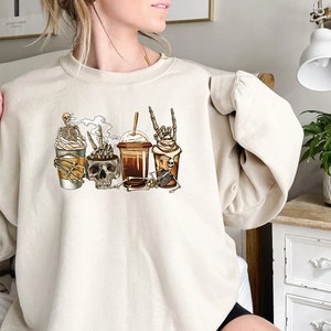 K&ouml;nnte beinhalten: Ein cremefarbener Pullover mit einem Aufdruck von vier Kaffeetassen mit Sch&auml;deln und Skeletten. Die Tassen sind mit Schlagsahne und Schokoladenso&szlig;e verziert.