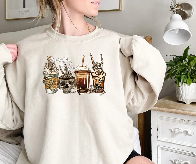 K&ouml;nnte beinhalten: Ein cremefarbener Pullover mit einem Aufdruck von vier Kaffeetassen mit Sch&auml;deln und Skeletten. Die Tassen sind mit Schlagsahne und Schokoladenso&szlig;e verziert.