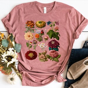 May include: A pink t-shirt with a floral print featuring a variety of flowers in shades of red, pink, yellow, and white.