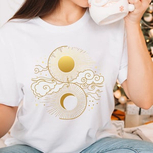 May include: White t-shirt with a gold sun and moon design. The design features a sun with rays, a crescent moon, and stylized clouds.