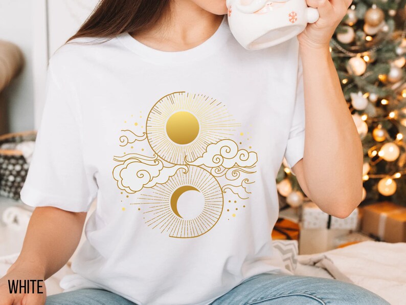 May include: White t-shirt with a gold sun and moon design. The design features a sun with rays, a crescent moon, and stylized clouds.