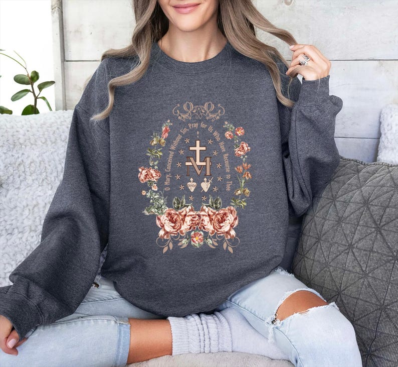 May include: A dark gray sweatshirt with a floral design and the text "Pray for Us Who Have Recourse to Thee O Mary Conceived Without Sin" in a circle around a cross and the letters "M" and "M".