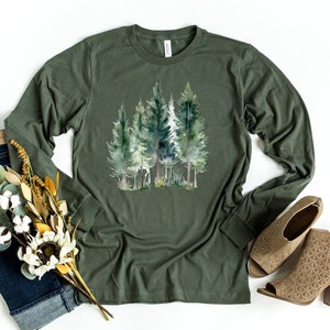 Puede incluir: Una camiseta de manga larga verde militar con un estampado de acuarela de una escena de bosque. El estampado presenta varios &aacute;rboles verdes con troncos marrones.
