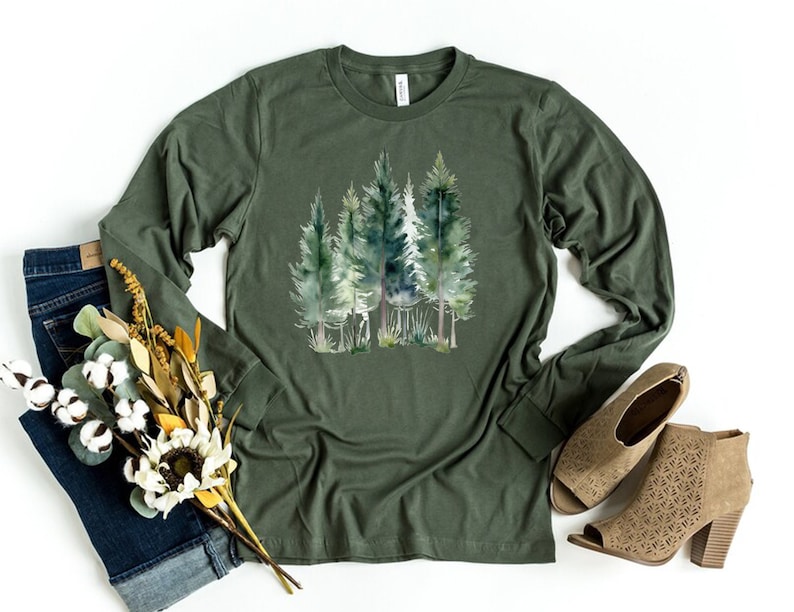 Puede incluir: Una camiseta de manga larga verde militar con un estampado de acuarela de una escena de bosque. El estampado presenta varios &aacute;rboles verdes con troncos marrones.