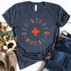 Puede incluir: Una camiseta azul marino con un dise&ntilde;o circular de colores que dice "Registered Nurse" con una cruz roja en el centro.