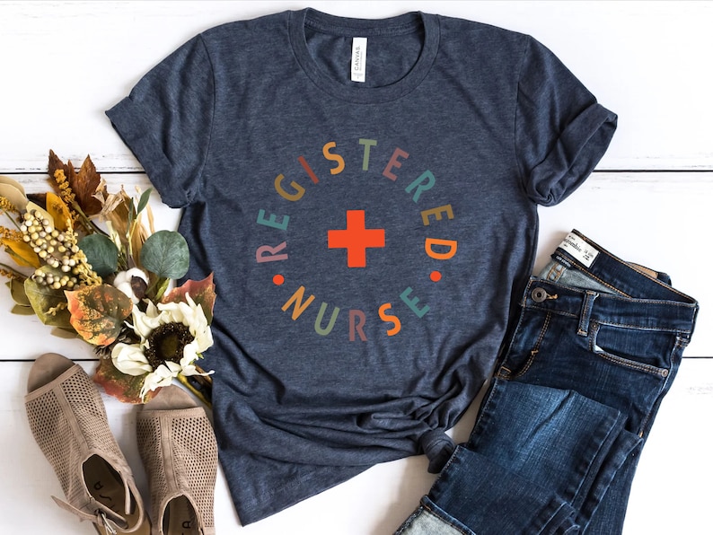 Puede incluir: Una camiseta azul marino con un dise&ntilde;o circular de colores que dice "Registered Nurse" con una cruz roja en el centro.