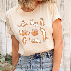 May include: Cream-colored t-shirt featuring a playful illustration of orange and white cats in various poses. The cats are depicted in a cartoon style, adding a whimsical touch to the design. The shirt is made of a soft material.