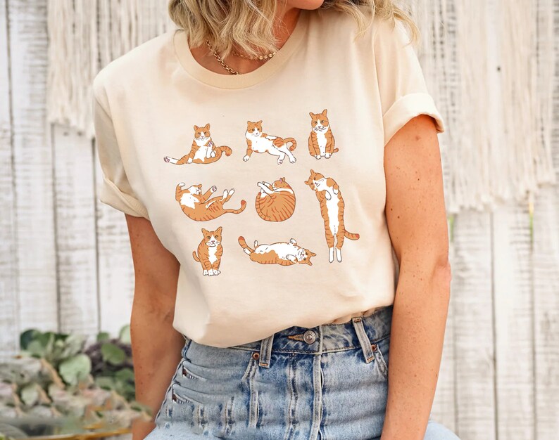 May include: Cream-colored t-shirt featuring a playful illustration of orange and white cats in various poses. The cats are depicted in a cartoon style, adding a whimsical touch to the design. The shirt is made of a soft material.