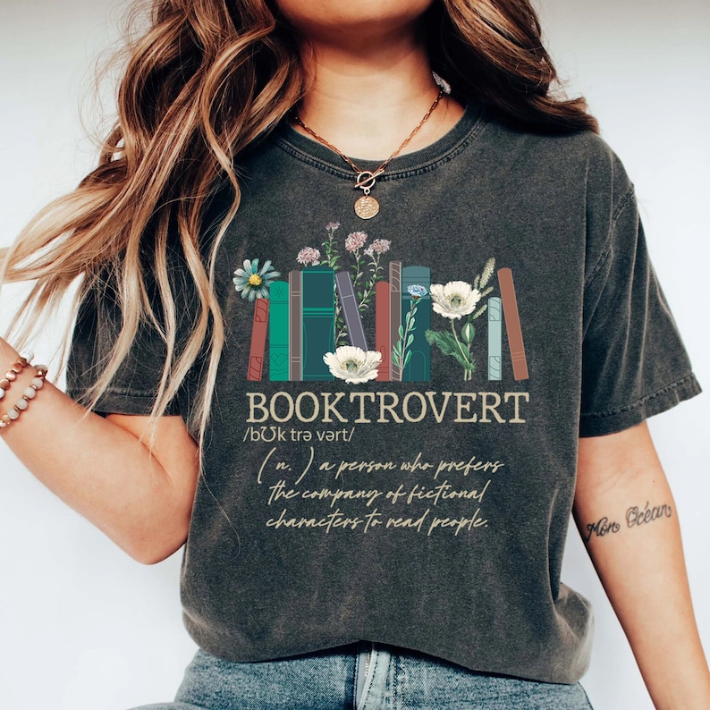 Puede incluir: Una camiseta gris oscuro con un gr&aacute;fico de libros apilados y flores. El texto "BOOKTROVERT /b&uuml;k trə vərt/ (n.) a person who prefers the company of fictional characters to read people." est&aacute; impreso en la camiseta.