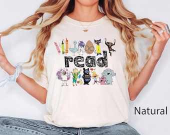 May include: A natural-colored short-sleeve t-shirt featuring a graphic print of various children's book characters surrounding the word "read" in black, scribbled-style lettering. The design includes colorful illustrations of crayons, animals, and figures.