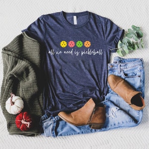 May include: A navy blue t-shirt with the text "all we need is pickleball" and four colorful pickleball paddles printed on the front.