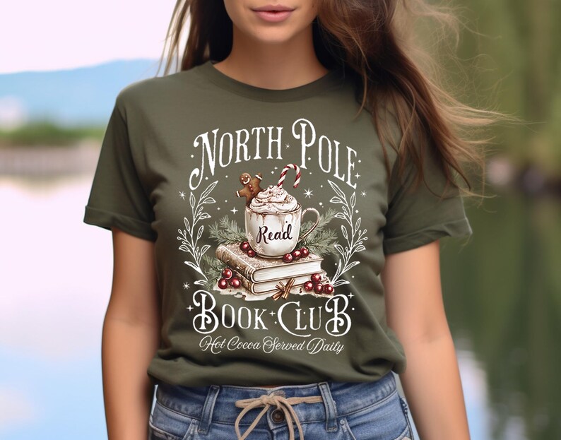 K&ouml;nnte beinhalten: Ein gr&uuml;nes T-Shirt mit einem wei&szlig;en Grafikdesign, auf dem "North Pole Book Club" steht, mit einer Tasse hei&szlig;er Schokolade und einem Stapel B&uuml;cher. Der Text lautet auch "Hot Cocoa Served Daily".