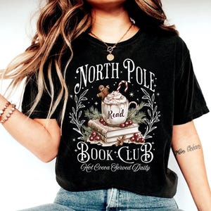 K&ouml;nnte beinhalten: Schwarzes T-Shirt mit einem wei&szlig;en Grafikdesign, das eine Tasse hei&szlig;e Schokolade mit Schlagsahne und einem Lebkuchenmann, einen Stapel B&uuml;cher und den Text "North Pole Book Club. Hot Cocoa Served Daily." zeigt.