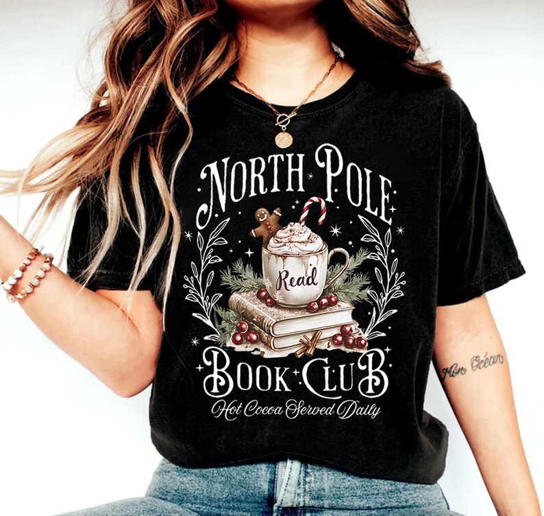 K&ouml;nnte beinhalten: Schwarzes T-Shirt mit einem wei&szlig;en Grafikdesign, das eine Tasse hei&szlig;e Schokolade mit Schlagsahne und einem Lebkuchenmann, einen Stapel B&uuml;cher und den Text "North Pole Book Club. Hot Cocoa Served Daily." zeigt.