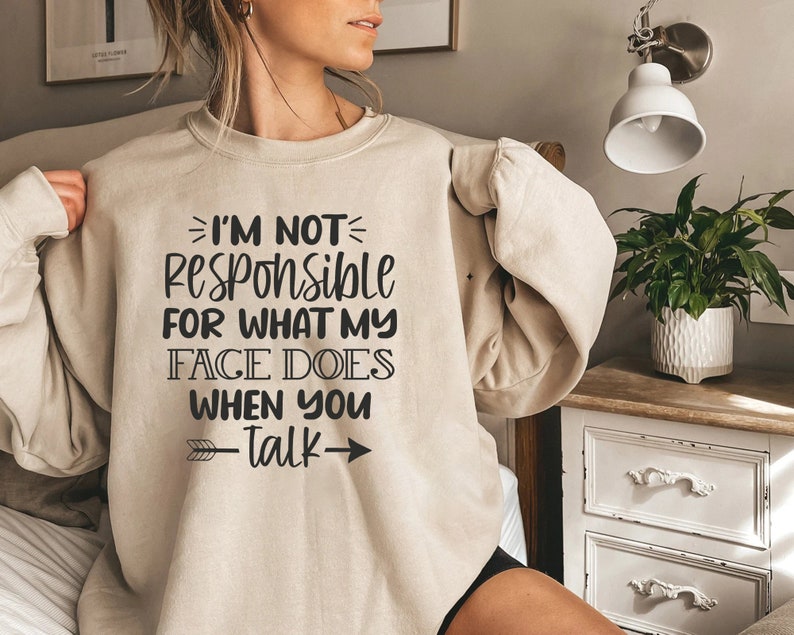 K&ouml;nnte beinhalten: Ein beiger Pullover mit dem Aufdruck "Ich bin nicht verantwortlich f&uuml;r das, was mein Gesicht tut, wenn du sprichst" in Schwarz.