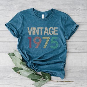 May include: A teal blue t-shirt with the text "Vintage 1975" in a retro font with colorful stripes. The numbers are in red, yellow, green, and blue.