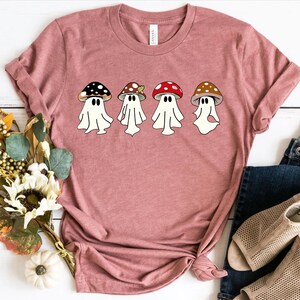 May include: A pink t-shirt featuring a design of four ghosts dressed as mushrooms. The ghosts are wearing different coloured mushroom caps, including red, brown, and black.