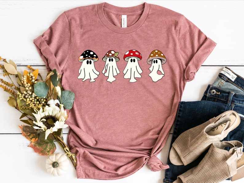 May include: A pink t-shirt featuring a design of four ghosts dressed as mushrooms. The ghosts are wearing different coloured mushroom caps, including red, brown, and black.