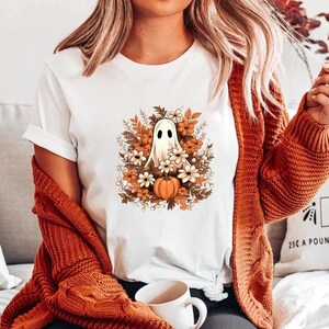 以下が含まれることがあります： 白いTシャツに、秋の葉とカボチャのリースの中に幽霊が描かれたグラフィックがプリントされています。