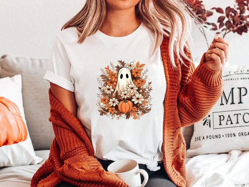 以下が含まれることがあります： 白いTシャツに、秋の葉とカボチャのリースの中に幽霊が描かれたグラフィックがプリントされています。