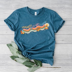 K&ouml;nnte beinhalten: Ein t&uuml;rkisfarbenes T-Shirt mit einem Regenbogen- und Blumenmotiv. Der Text auf dem Shirt lautet "Progress is not linear."