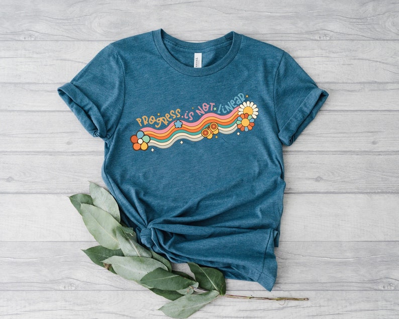 K&ouml;nnte beinhalten: Ein t&uuml;rkisfarbenes T-Shirt mit einem Regenbogen- und Blumenmotiv. Der Text auf dem Shirt lautet "Progress is not linear."
