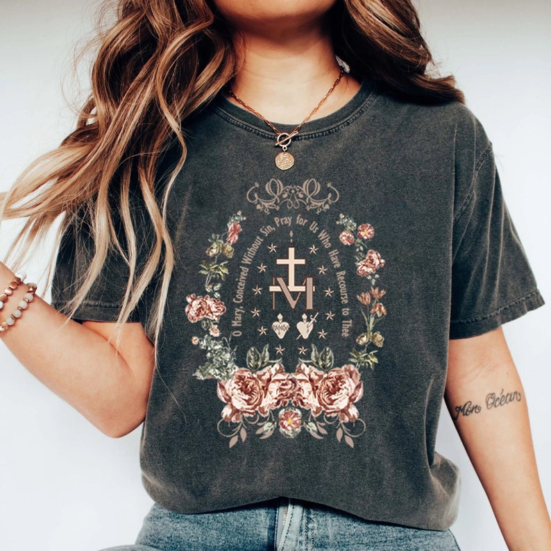 Op de afbeelding: Een donkergrijs T-shirt met een bloemenmotief en de tekst "O Maria, ontvangen zonder zonde, bid voor ons die onze toevlucht tot u nemen." Het ontwerp bevat een kruis, een hart en een kroon.