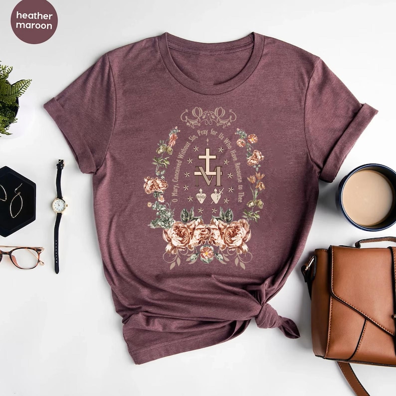 Op de afbeelding: Een heide-marron t-shirt met een bloemenmotief en de tekst "O Maria, ontvangen zonder zonde, bid voor ons die onze toevlucht tot u nemen" in een cirkel rond een kruis en de letters "M" en "V".