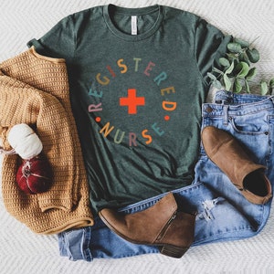 Puede incluir: Una camiseta verde oscuro con un gr&aacute;fico circular de colores que dice "REGISTERED NURSE" con una cruz roja en el centro.
