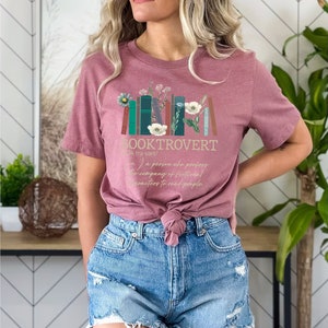 Puede incluir: Una camiseta rosa con un dise&ntilde;o gr&aacute;fico de una pila de libros con flores y el texto "BOOKTROVERT (bk tra vart/) a person who prefers the company of fictional characters to read people."