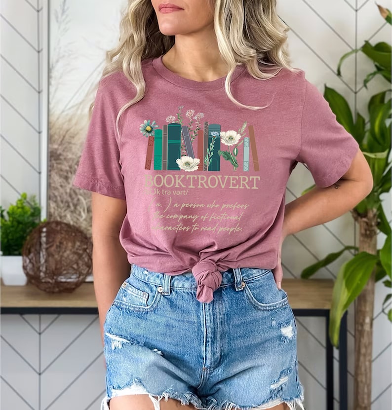 Puede incluir: Una camiseta rosa con un dise&ntilde;o gr&aacute;fico de una pila de libros con flores y el texto "BOOKTROVERT (bk tra vart/) a person who prefers the company of fictional characters to read people."
