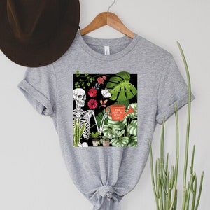 May include: Gray t-shirt with a graphic of a skeleton holding a watering can with the text "You make me feel alive". The graphic also includes plants and flowers.