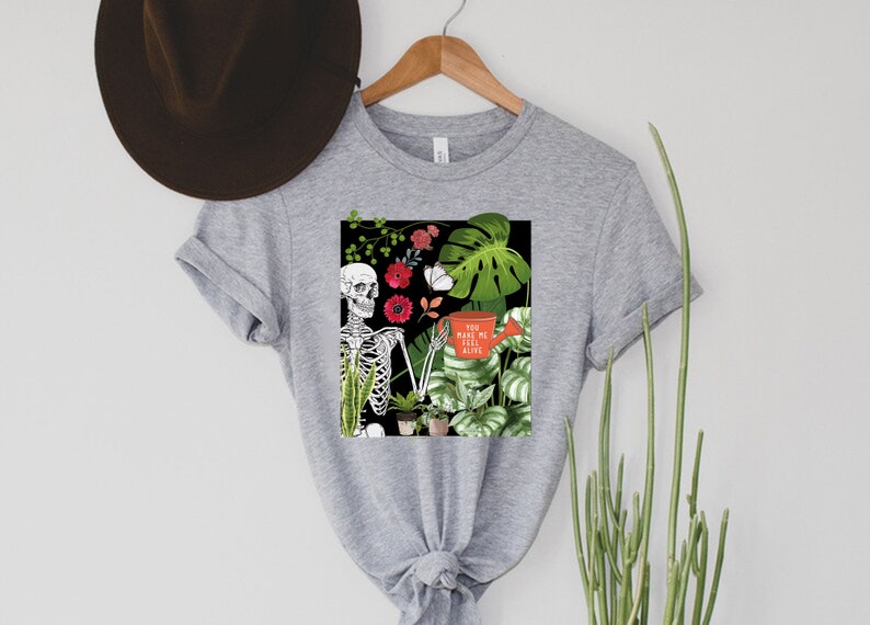 May include: Gray t-shirt with a graphic of a skeleton holding a watering can with the text "You make me feel alive". The graphic also includes plants and flowers.
