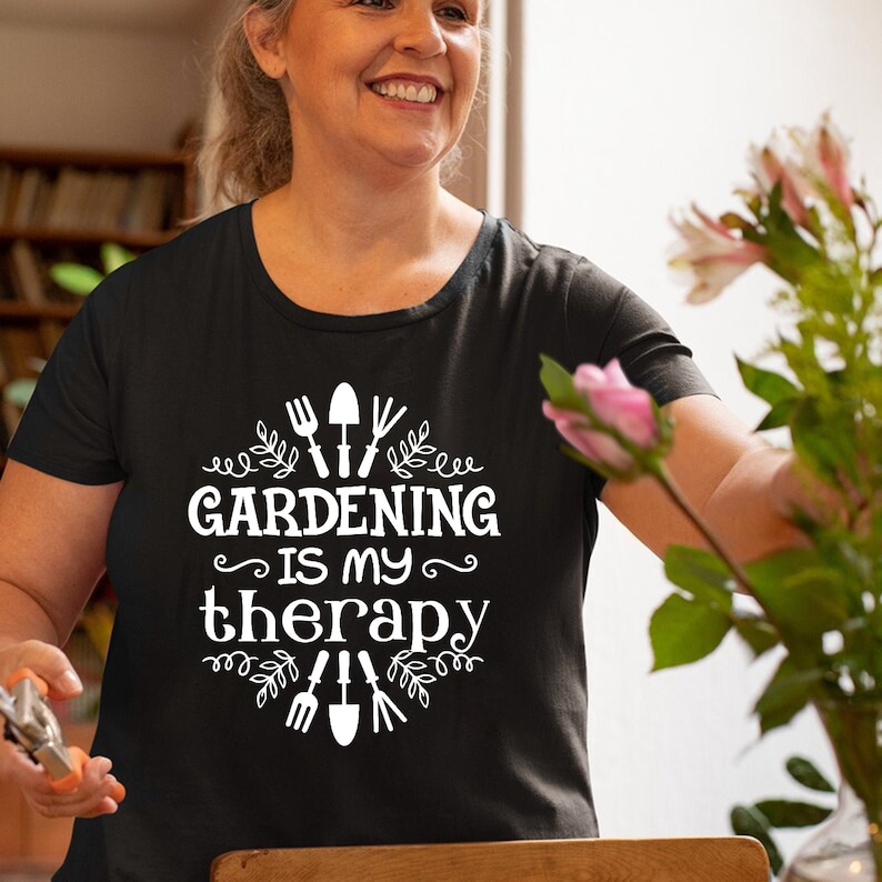 May include: A black t-shirt with white text that reads "Gardening is my therapy" surrounded by gardening tools and leaves.