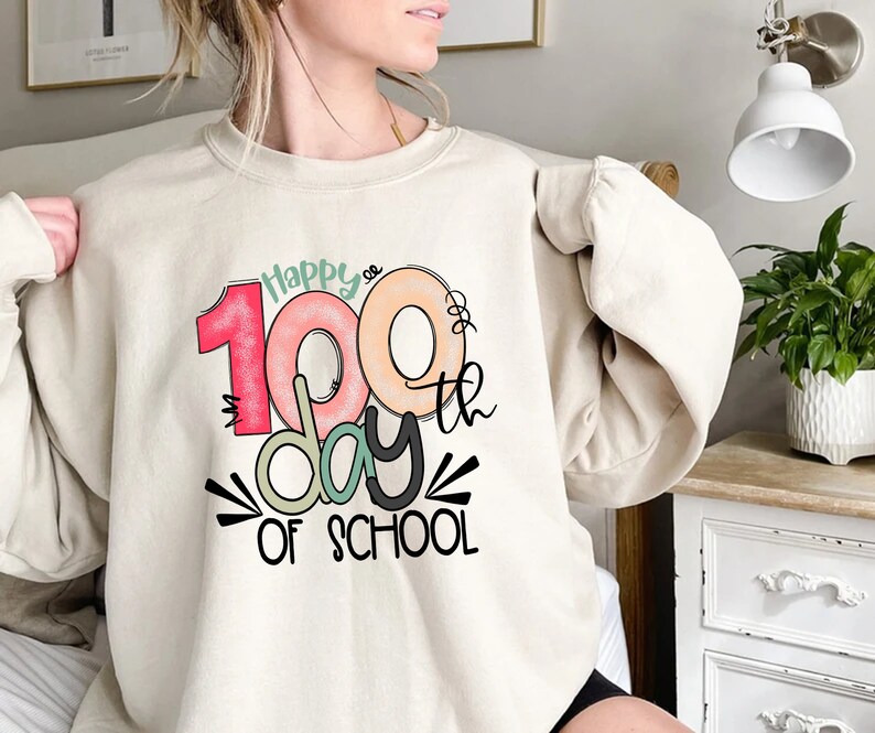 May include: A beige sweatshirt with a graphic design that says "Happy 100th day of school". The number 100 is stylized to look like a donut.