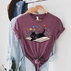 May include: A maroon t-shirt with a black cat reading a book surrounded by colorful flowers. The cat is looking at the book and has a curious expression.