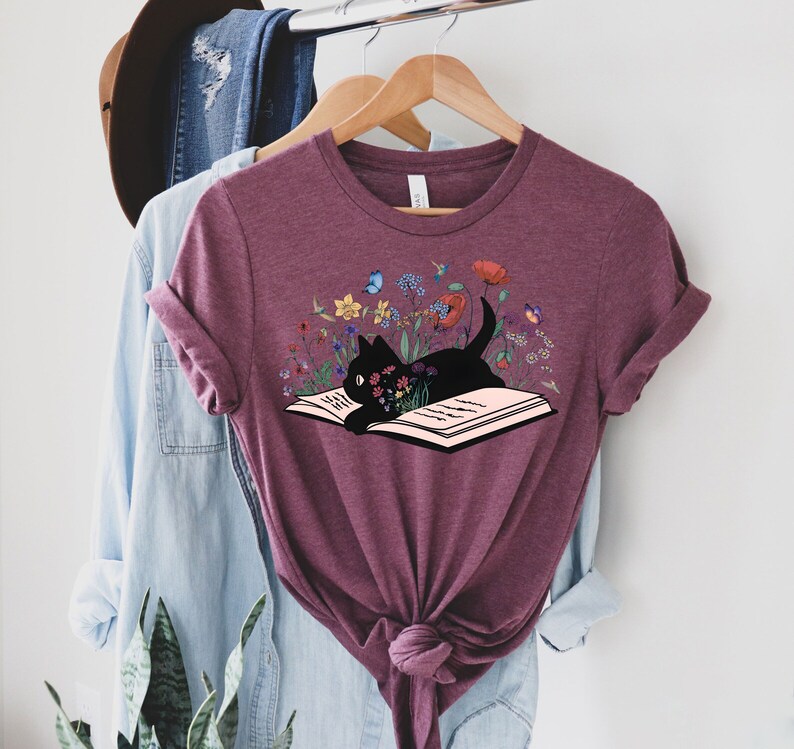 May include: A maroon t-shirt with a black cat reading a book surrounded by colorful flowers. The cat is looking at the book and has a curious expression.