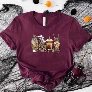 K&ouml;nnte beinhalten: Ein maroonfarbenes T-Shirt mit einem Motiv von drei Kaffeebetr&auml;nken mit Skeletten und einem Sch&auml;del. Die Kaffeebetr&auml;nke sind mit Schlagsahne und Schokolade verziert.