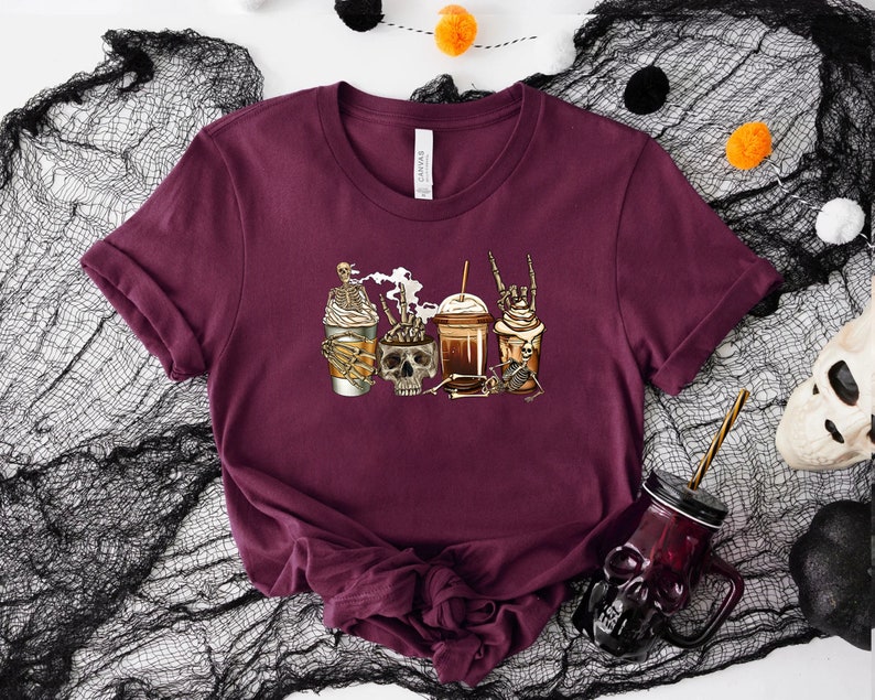 K&ouml;nnte beinhalten: Ein maroonfarbenes T-Shirt mit einem Motiv von drei Kaffeebetr&auml;nken mit Skeletten und einem Sch&auml;del. Die Kaffeebetr&auml;nke sind mit Schlagsahne und Schokolade verziert.