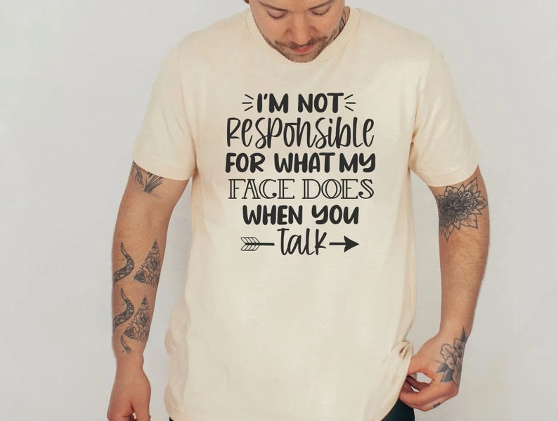 K&ouml;nnte beinhalten: Ein beiges T-Shirt mit einem schwarzen Aufdruck, der "Ich bin nicht verantwortlich f&uuml;r das, was mein Gesicht tut, wenn du sprichst" lautet, mit einem Pfeil, der nach rechts zeigt.