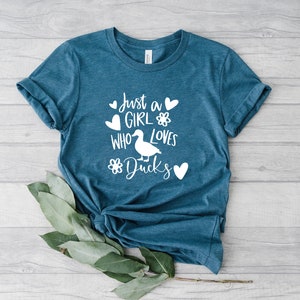 May include: Teal-colored t-shirt with the text "Just a GIRL who loves Ducks" in white. The design includes a duck illustration, hearts, and flower accents. The shirt is made of soft material.