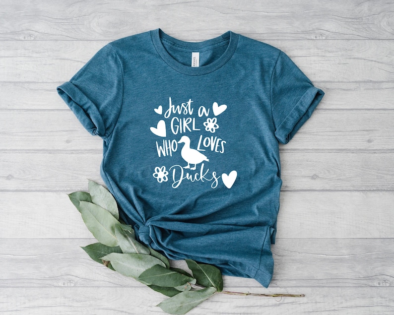 May include: Teal-colored t-shirt with the text "Just a GIRL who loves Ducks" in white. The design includes a duck illustration, hearts, and flower accents. The shirt is made of soft material.