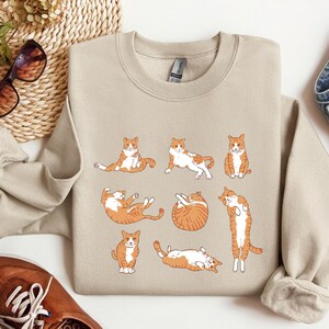 K&ouml;nnte beinhalten: Ein beiger Pullover mit einem grafischen Druck von neun orangefarbenen und wei&szlig;en Katzen in verschiedenen Posen. Die Katzen sind alle nach vorne gerichtet und haben unterschiedliche Gesichtsausdr&uuml;cke.