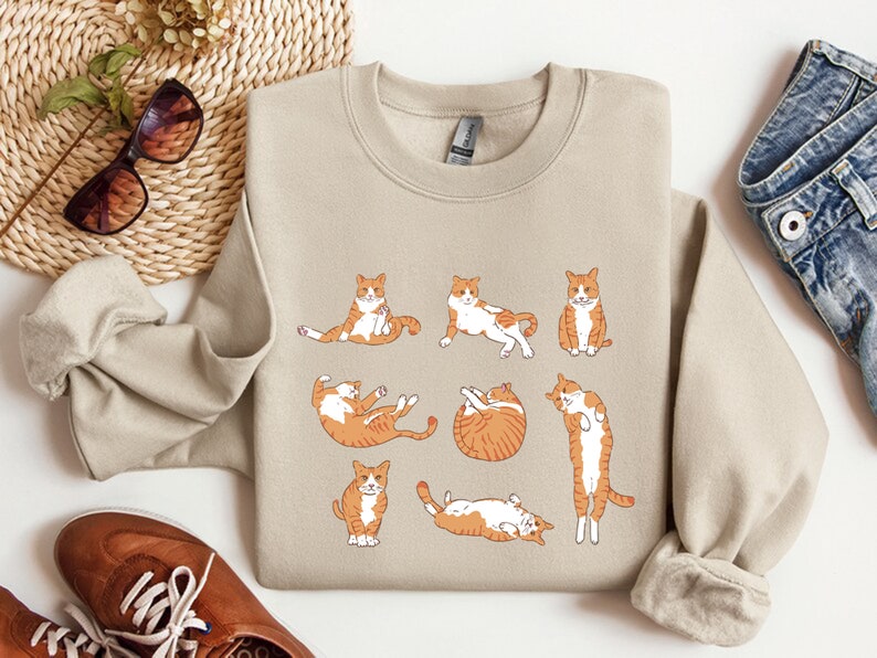 K&ouml;nnte beinhalten: Ein beiger Pullover mit einem grafischen Druck von neun orangefarbenen und wei&szlig;en Katzen in verschiedenen Posen. Die Katzen sind alle nach vorne gerichtet und haben unterschiedliche Gesichtsausdr&uuml;cke.